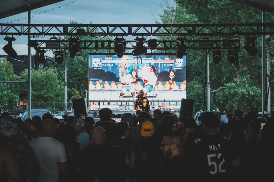 DJ performing in front of a screen showing a retro fighting game, crowd in silhouette