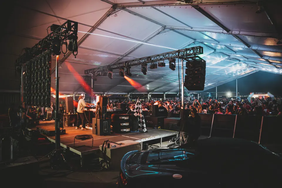 Side view of the stage with a DJ, a checkered flag, and a large crowd under the lit tent